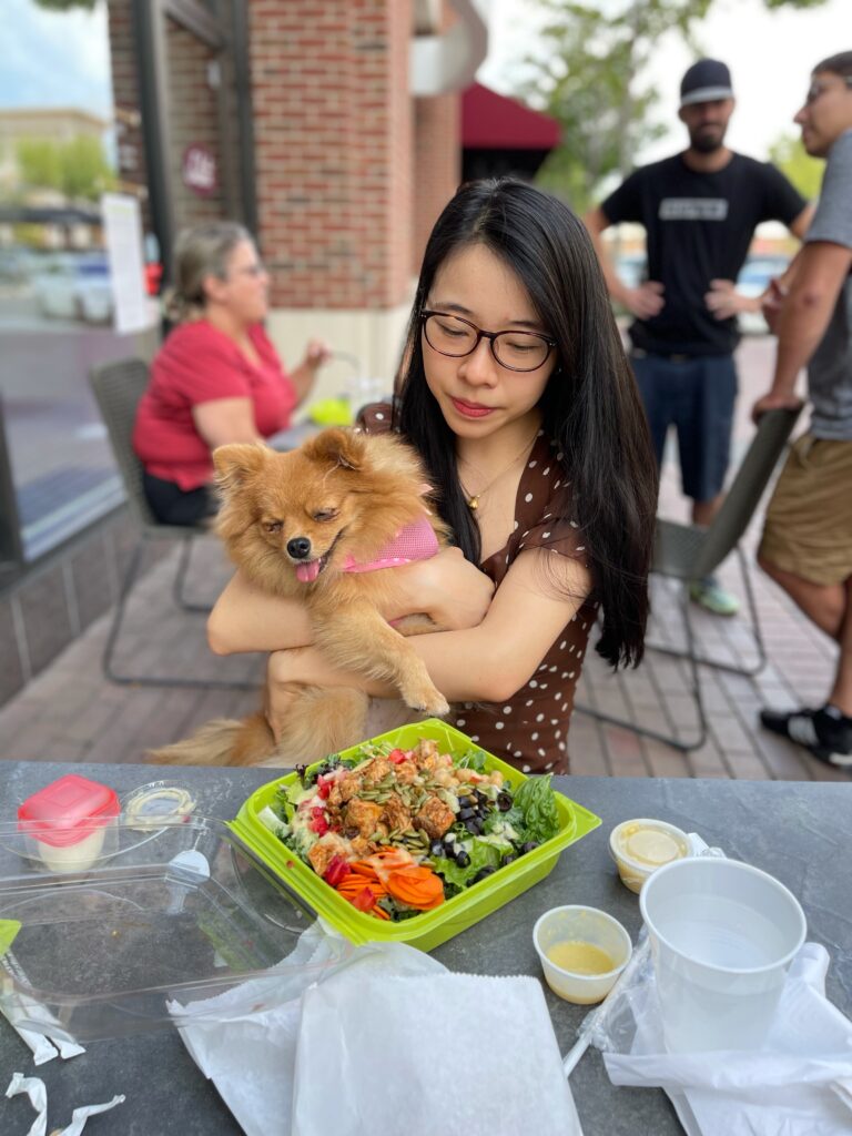 Vivian, a girl in brown polka dot dress, holds her pomeranian puppy with her lunch, salata salad
